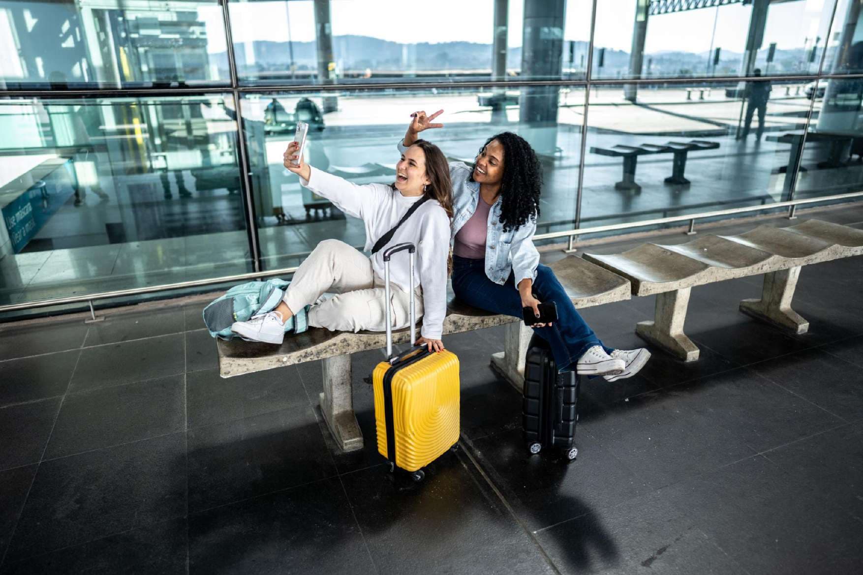 Two friends taking a selfie on an airport bench with suitcases